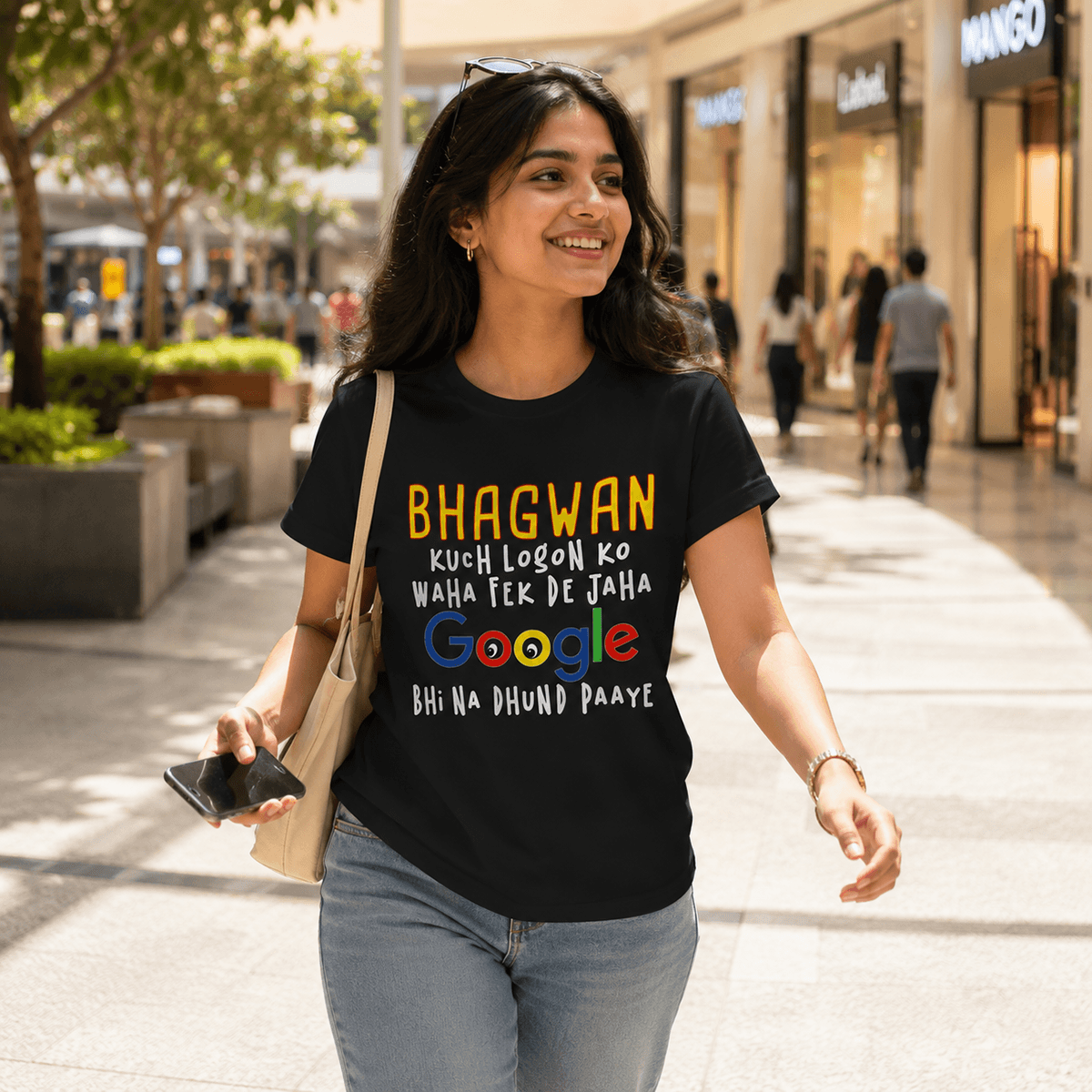 Smiling woman walking outdoors in a shopping street, wearing a black t-shirt with a colorful Google logo and Hindi text, holding a phone and a beige bag.